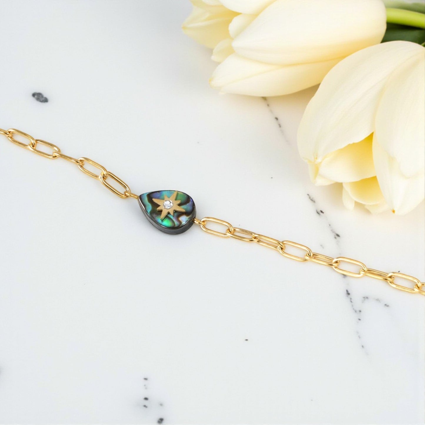 Gold bracelet with a heart-shaped abalone shell charm on a marble surface with white tulips.