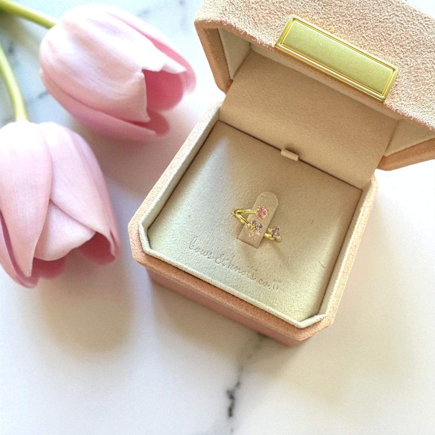 Gold ring with pink gemstone in a jewelry box with pink tulips on a marble surface