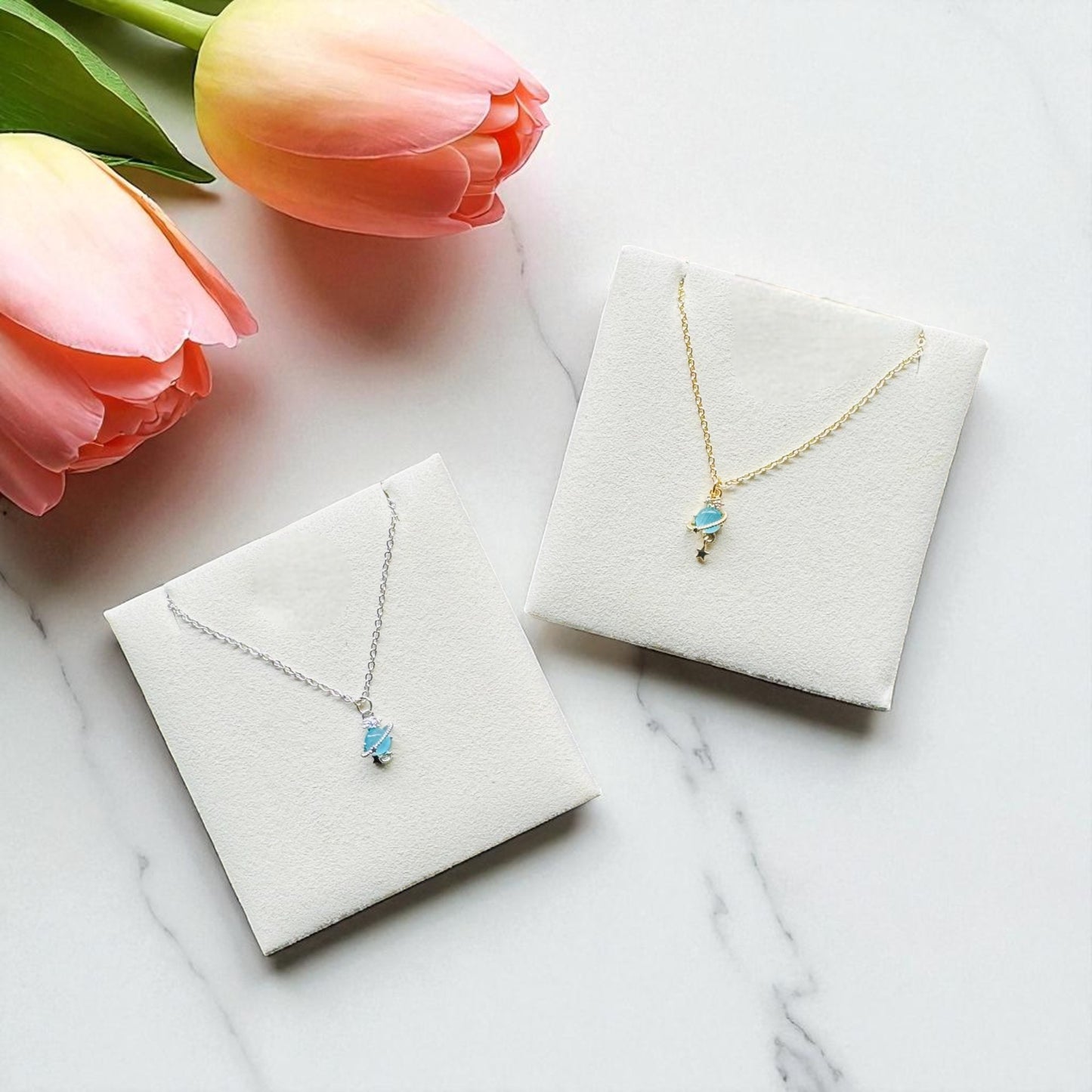 Two necklaces in display cases with pink tulips on a marble surface