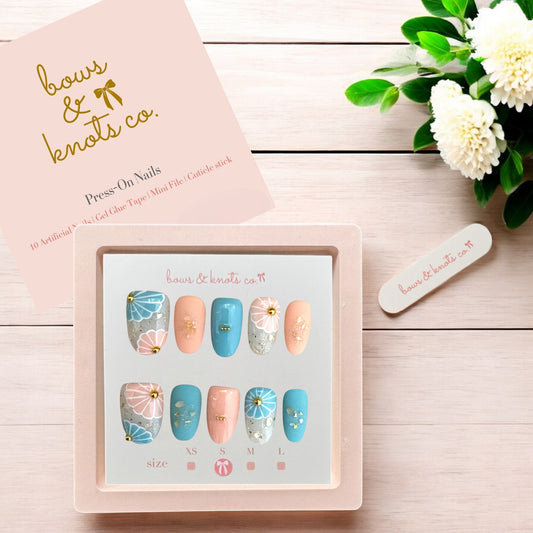 Set of press-on nails with decorative designs in a box on a wooden surface with flowers.