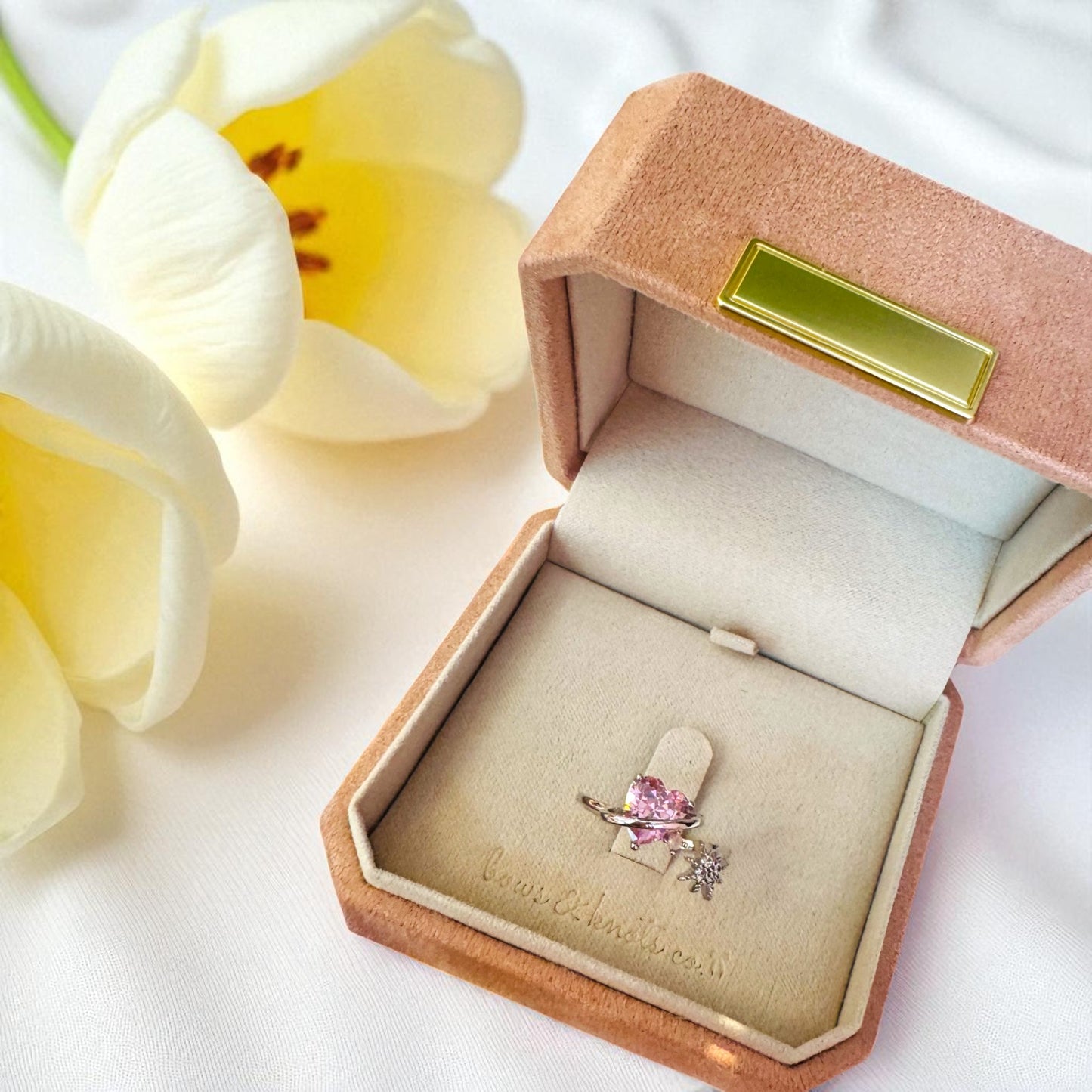 Jewelry box with a ring featuring a pink gemstone and diamond, surrounded by white flowers.