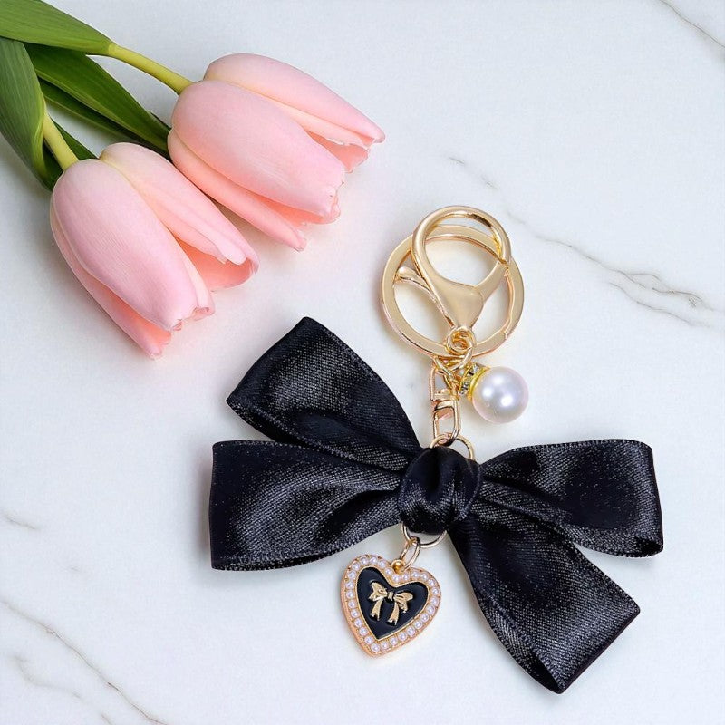 Black bow keychain with heart charm and pearl on a white surface with pink tulips.