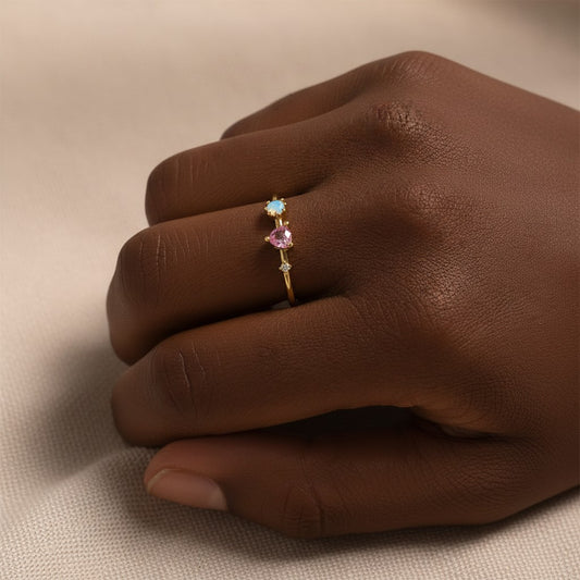 A close-up of a hand wearing a delicate gold ring with three small heart-shaped gemstones in light blue, pink, and yellow tones, resting on a neutral beige fabric background.