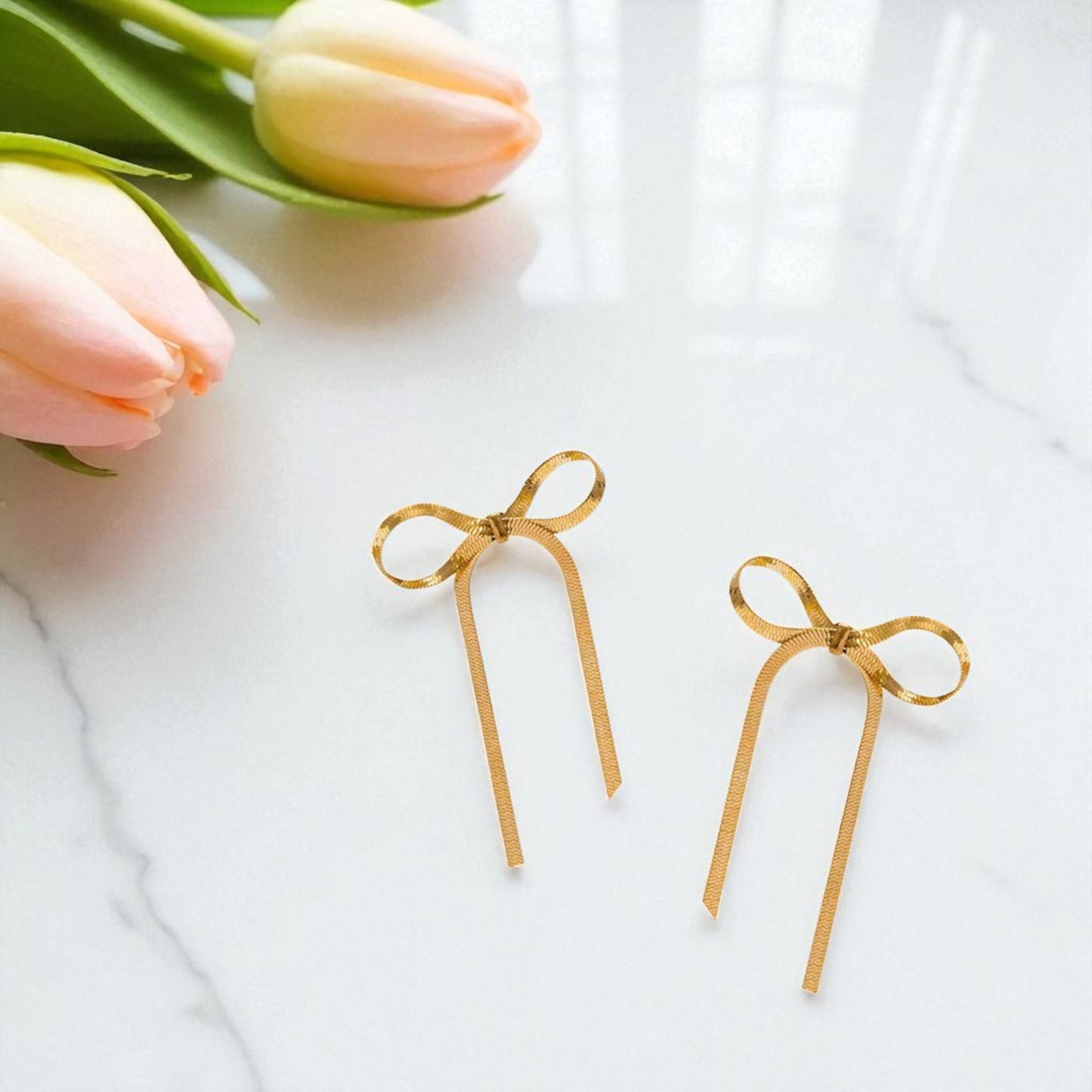 Gold bow-shaped earrings on a white surface with pink tulips.