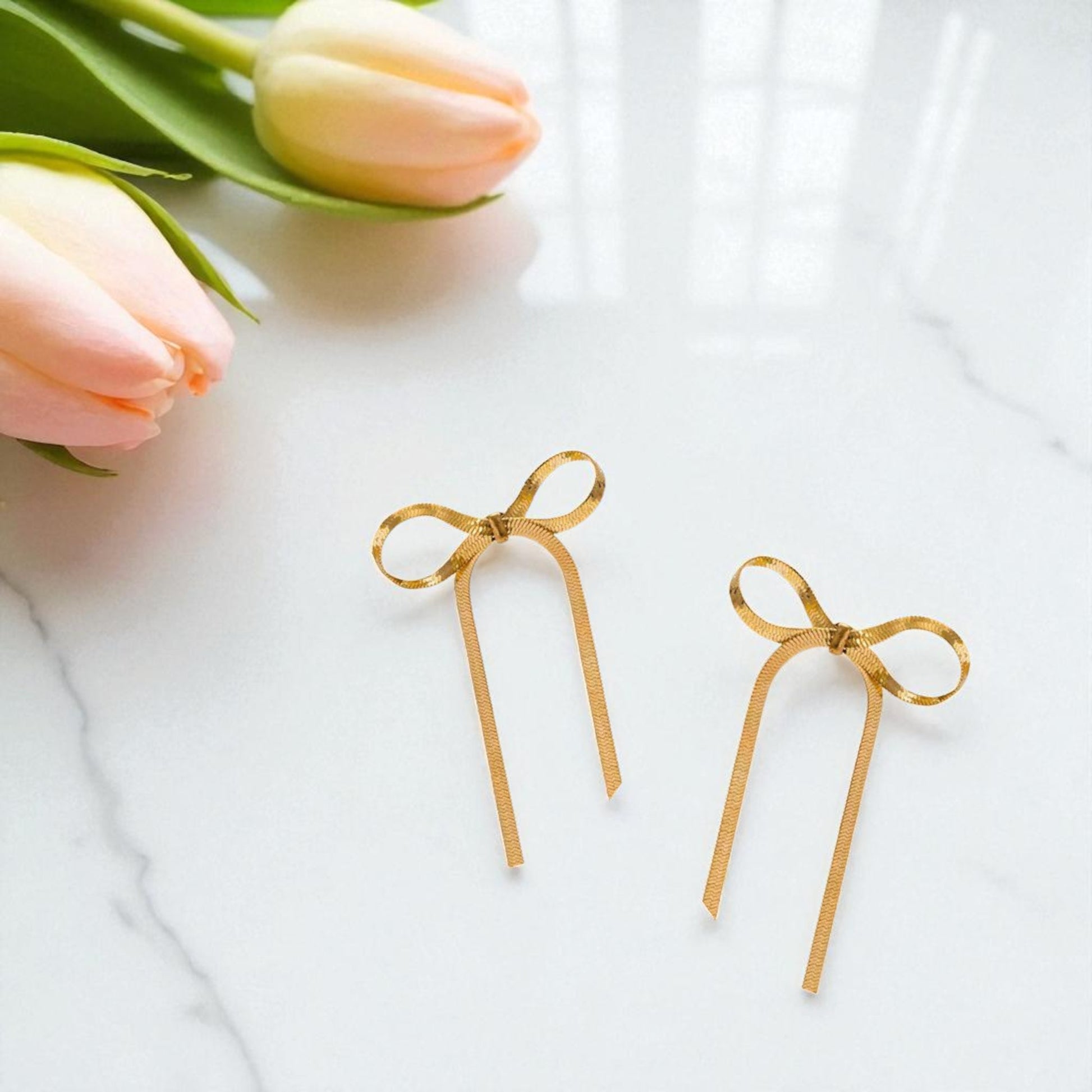Gold bow-shaped earrings on a white surface with pink tulips.