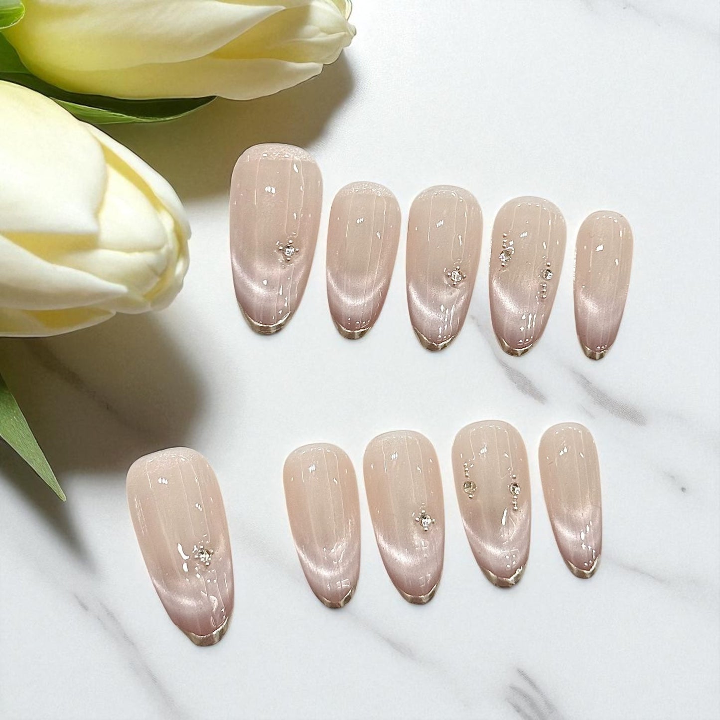 Set of nude-colored nail polish samples with decorative crystals on a white surface with tulips.