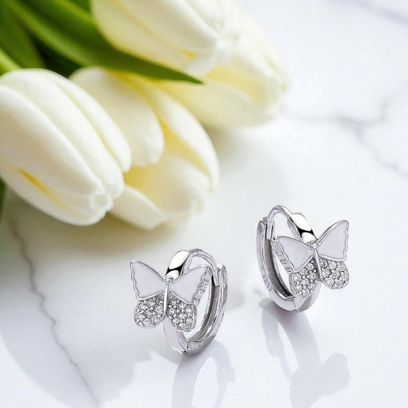 Butterfly-shaped silver earrings with white stones on a white surface with tulips in the background.