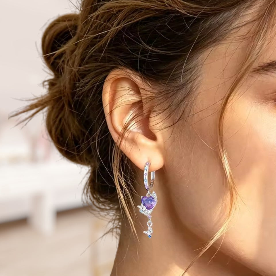 A close-up of a woman's ear wearing a silver huggie hoop earring with a dangling heart-shaped purple gemstone and clear crystal accents.