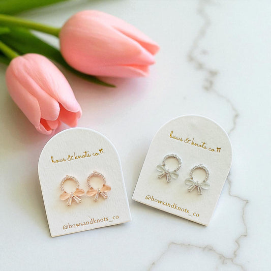 Pair of bow-shaped earrings on branded display cards with pink tulips in the background.