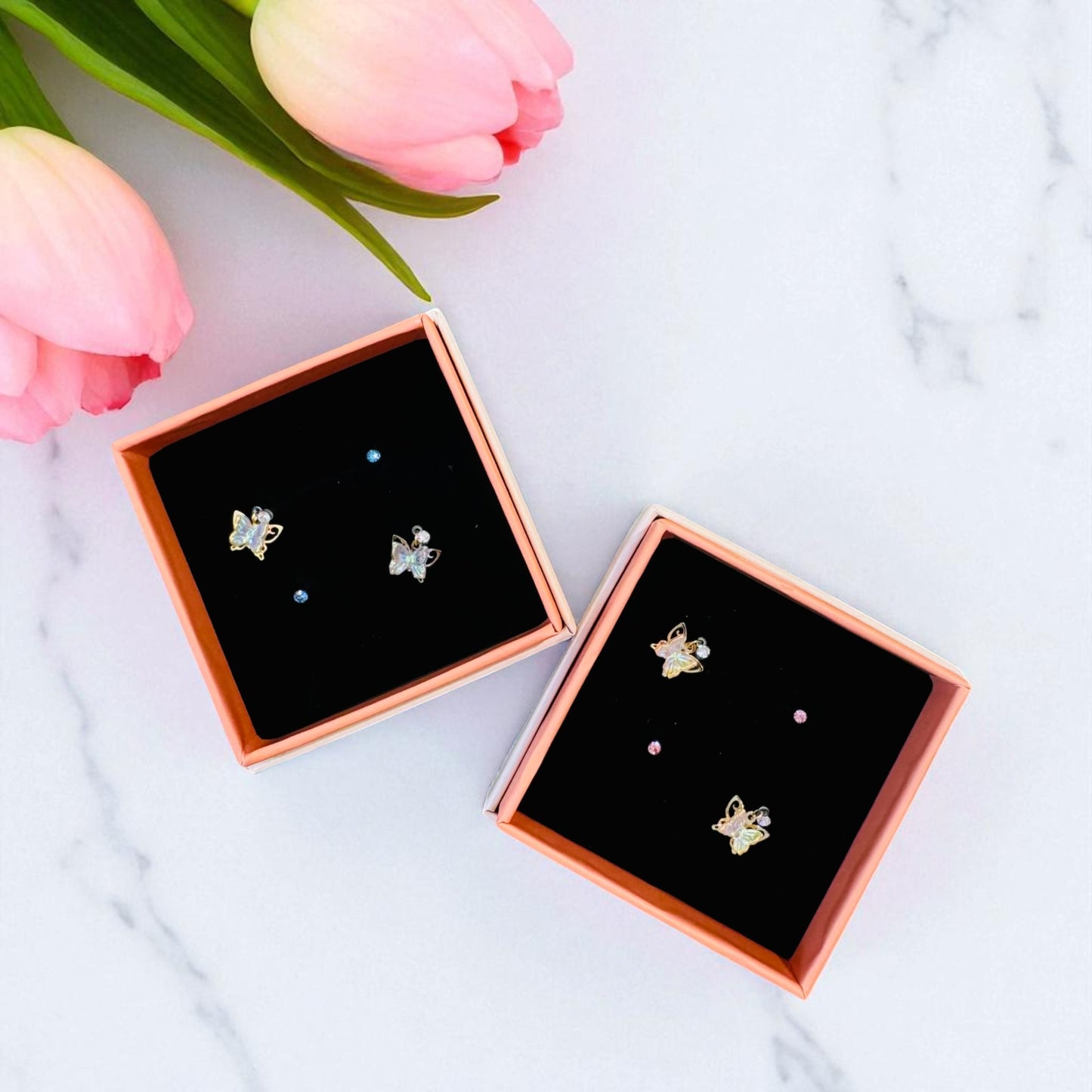 Butterfly-shaped earrings in rose gold and silver in a box with pink tulips on a marble background
