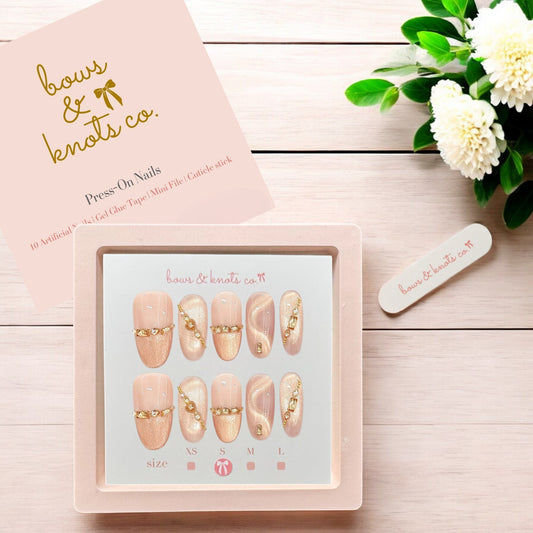 Set of press-on nails with decorative elements in a box, branded 'bows & knots co', on a wooden surface with flowers.