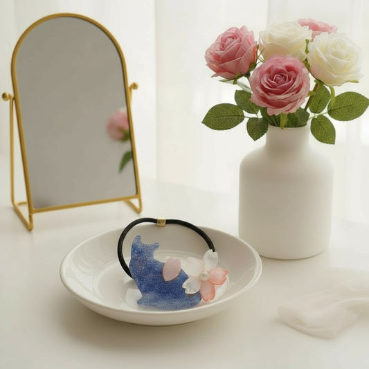 Decorative hair tie with a cat and cherry blossom flower design on a white dish, next to a vase of pink and white roses and a small mirror.