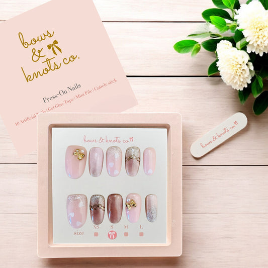 Set of press-on nails with a pink and gold design, placed on a wooden surface with a floral arrangement.