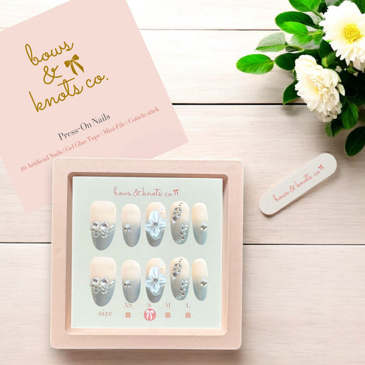 Set of press-on nails with decorative elements on a wooden surface, accompanied by a pink box and flowers.