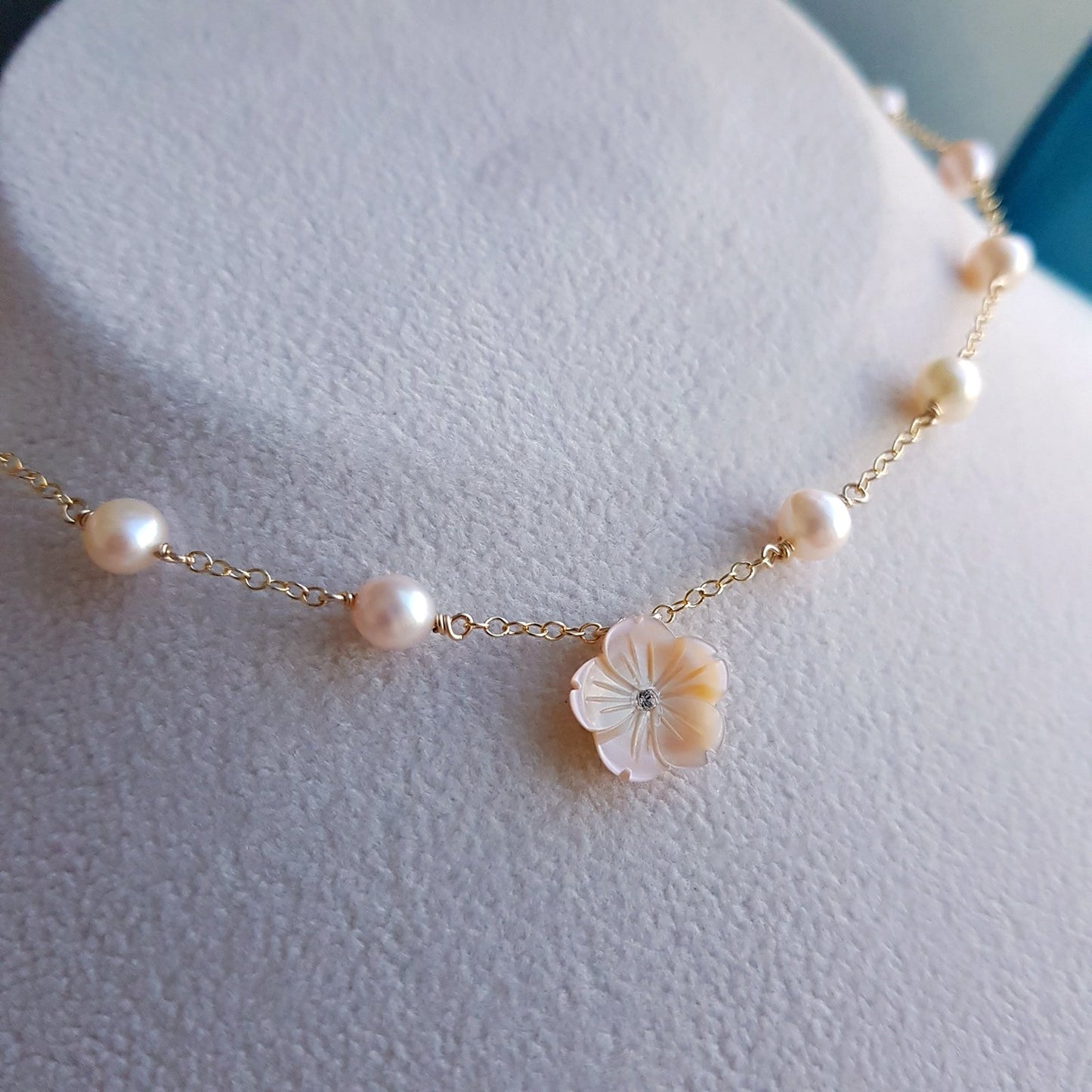 A delicate gold chain necklace with scattered white freshwater pearls and a central iridescent shell flower charm, displayed on a textured white jewelry bust.