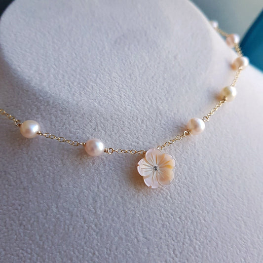 A delicate gold chain necklace with scattered white freshwater pearls and a central iridescent shell flower charm, displayed on a textured white jewelry bust.