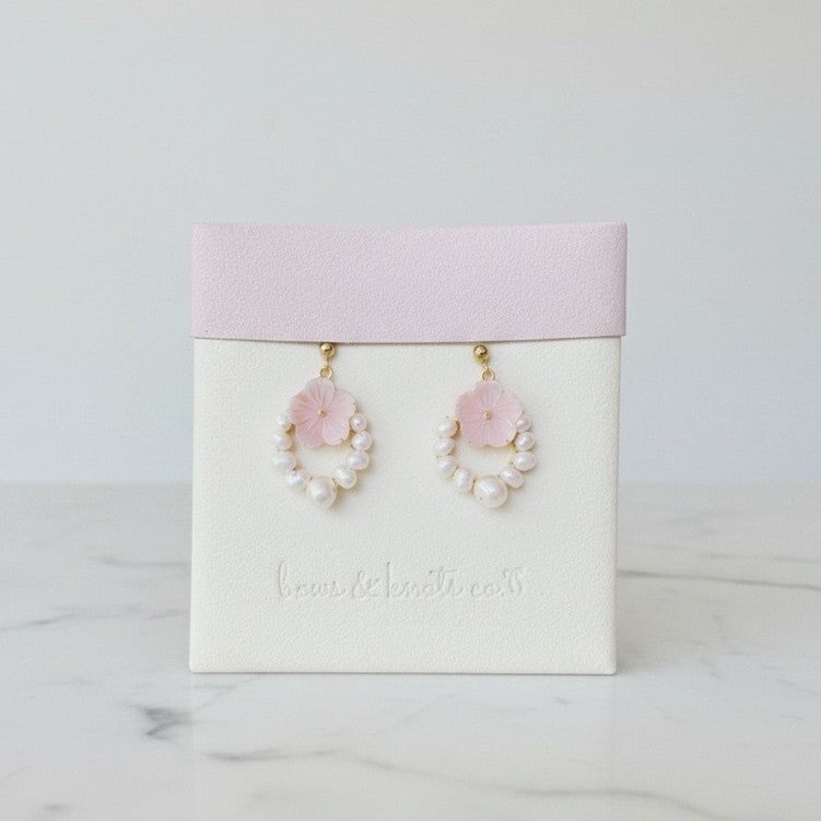A pair of dangling earrings displayed on a white jewelry card with a pale pink top band. Each earring has a small pink flower above a ring of white faux pearls, with gold-tone hooks.