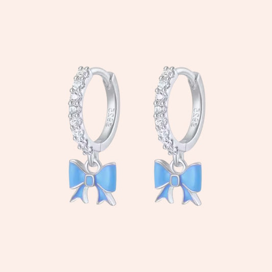 Pair of silver hoop earrings with blue bow charms on a light pink background