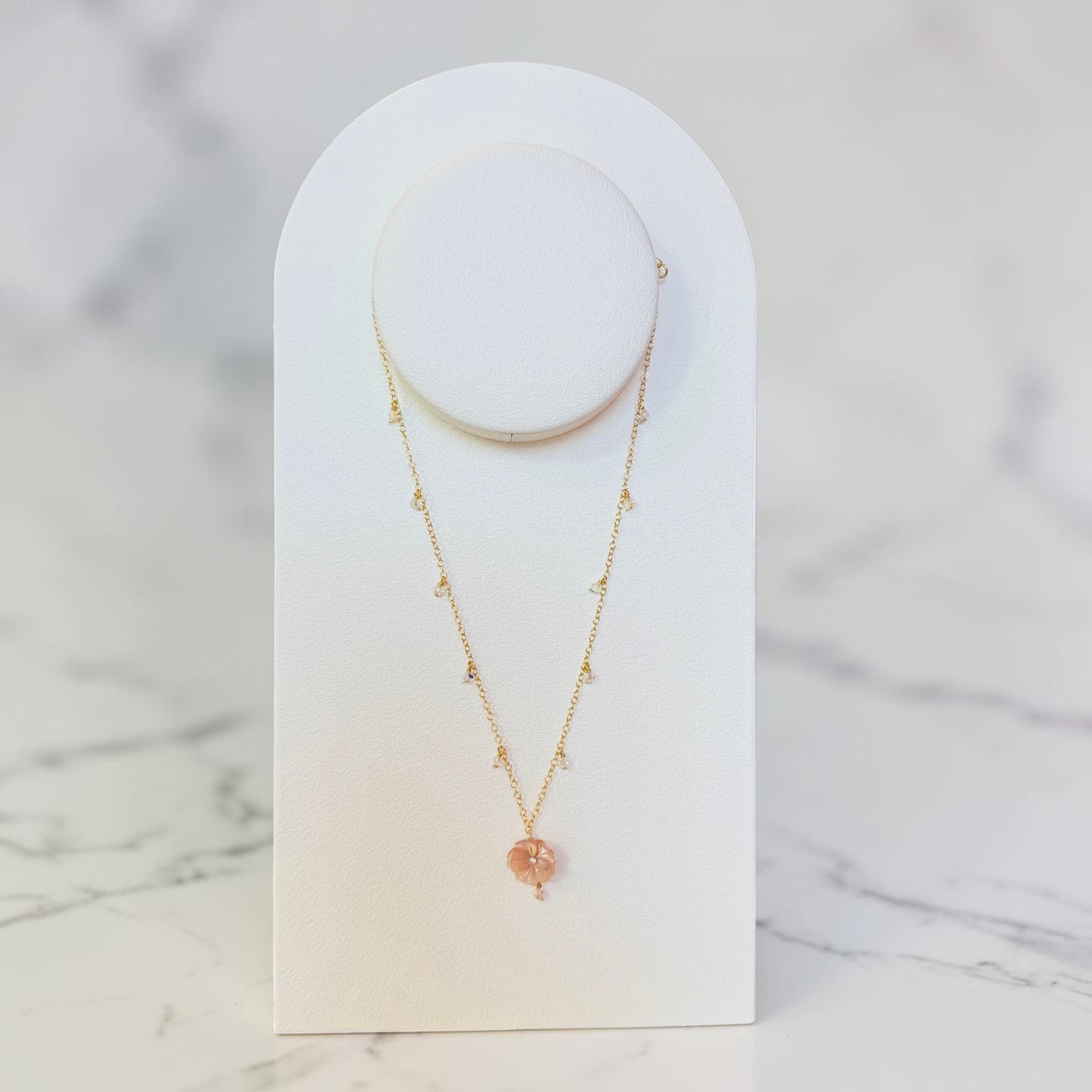 A delicate gold necklace with a small peach-pink heart-shaped crystal pendant, displayed on a white arched jewelry stand against a light marble background.