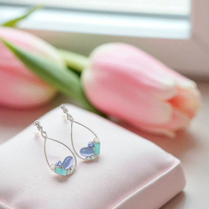 Teardrop-shaped earrings with gemstones on a pink surface with tulips in the background