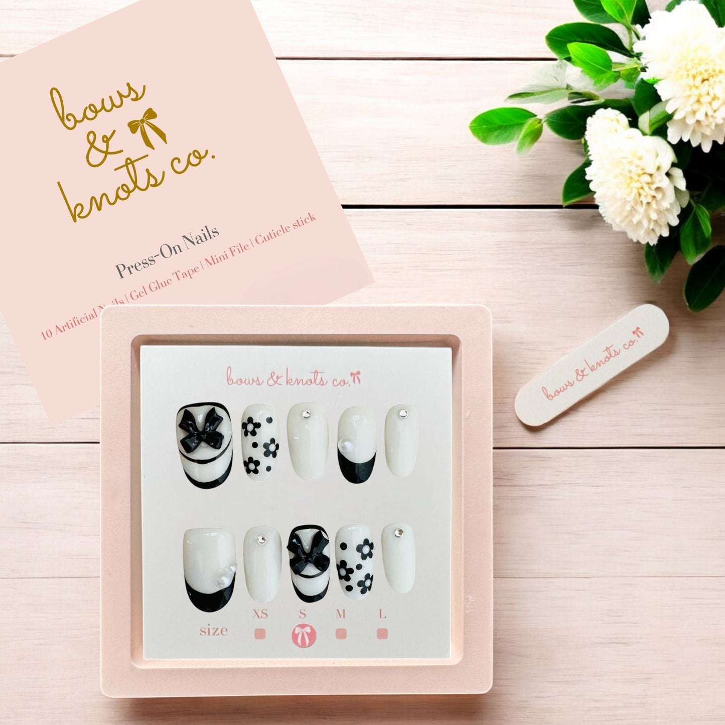 Set of press-on nails with a 'bows & knots co.' box on a wooden surface with flowers.