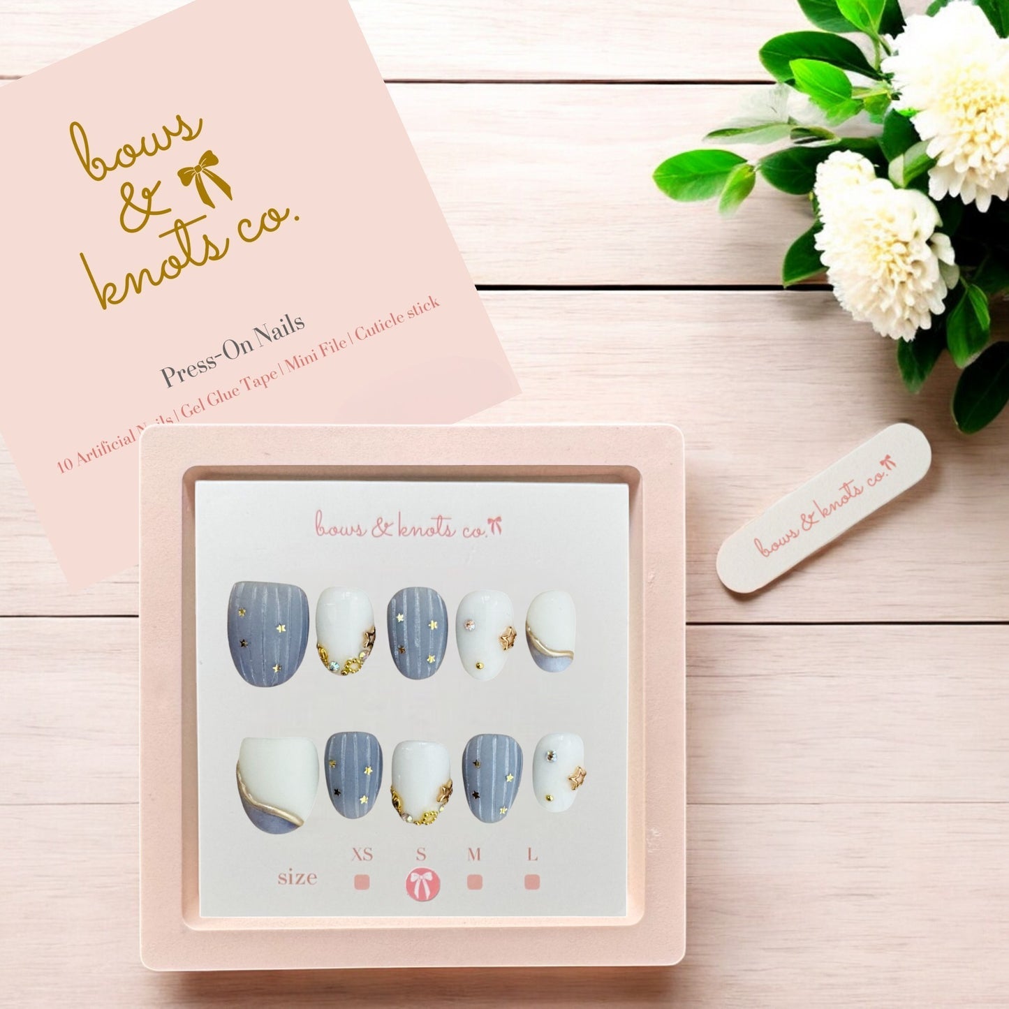 Set of press-on nails in a box with 'bows & knots co.' branding on a wooden surface with flowers.