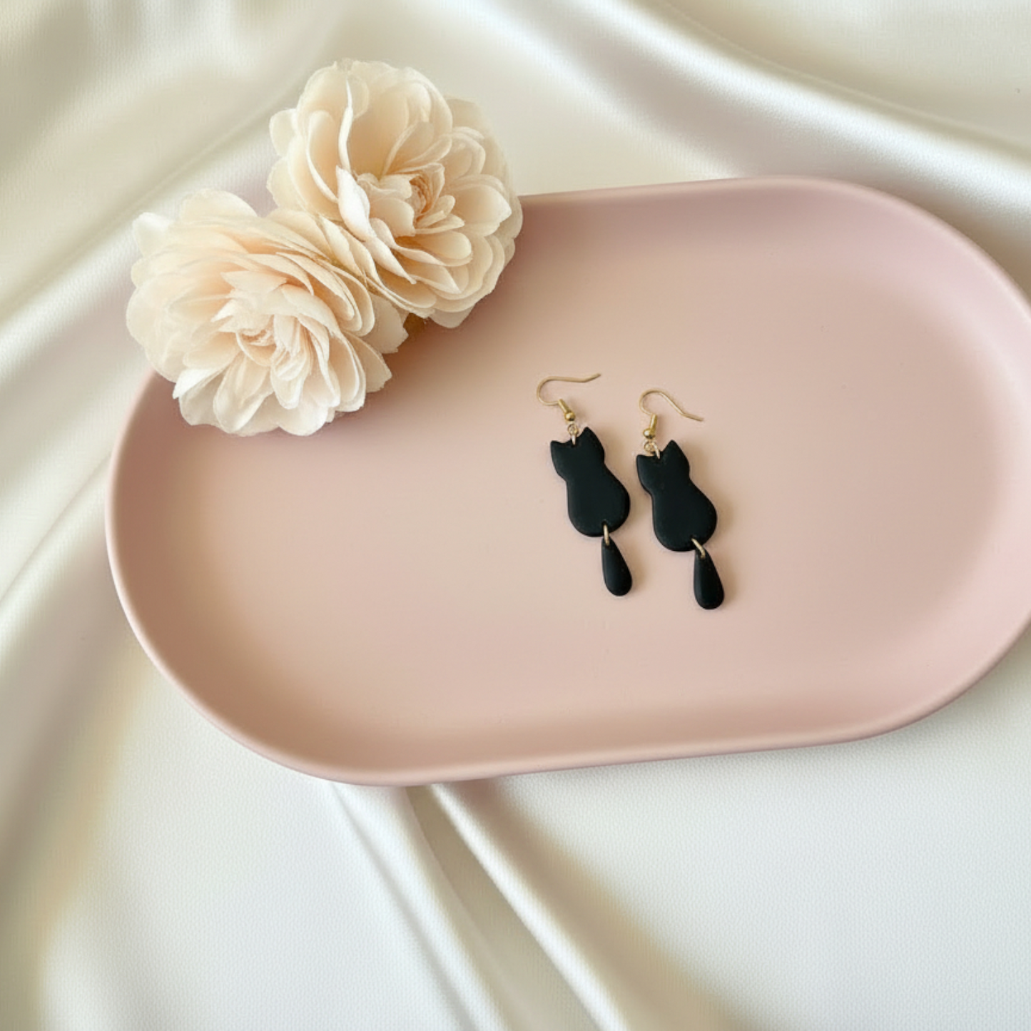 Black cat-shaped earrings on a pink tray with a beige flower