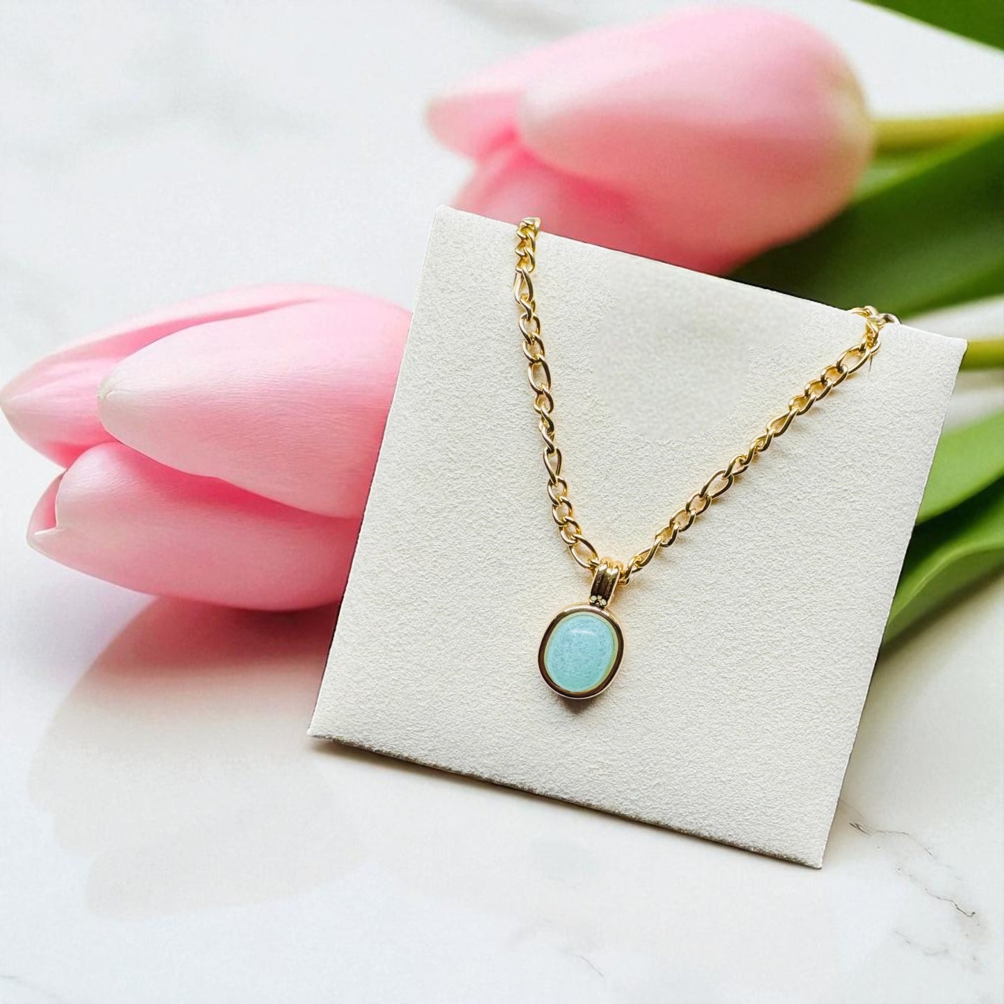 Gold necklace with a turquoise pendant on a white card with pink tulips in the background