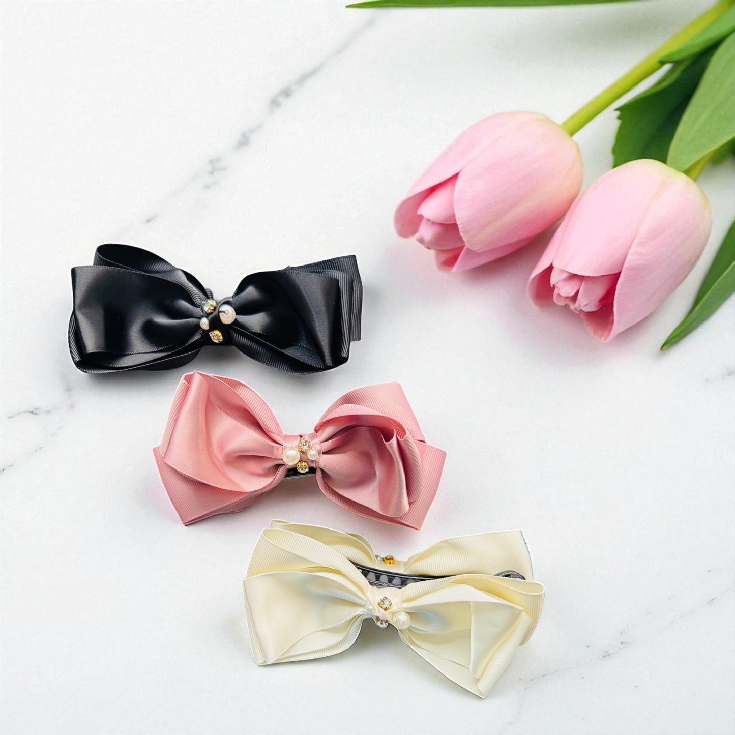 Three decorative bows in black, pink, and beige on a marble surface with pink tulips.