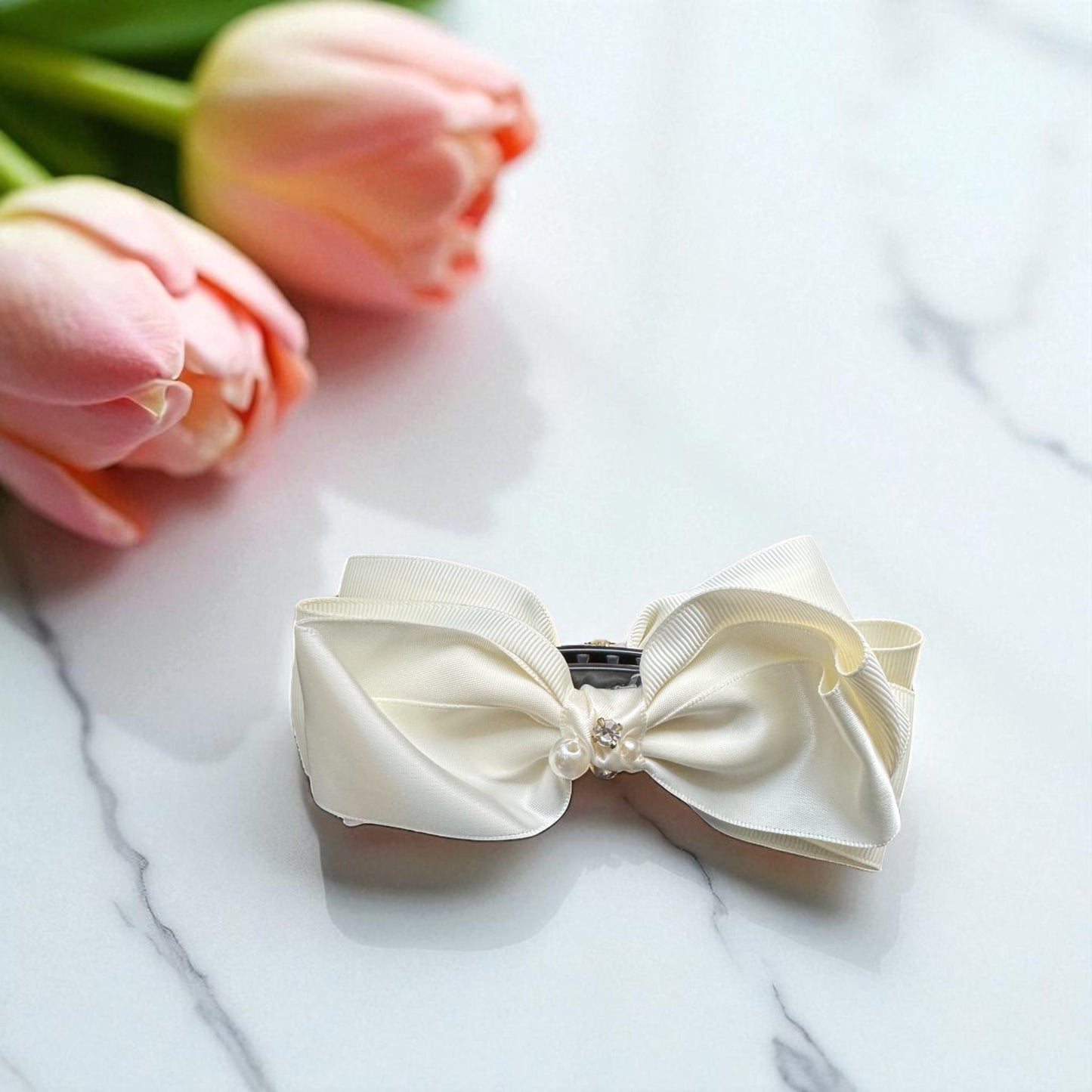 Cream-colored bow hair clip on a marble surface with pink tulips in the background