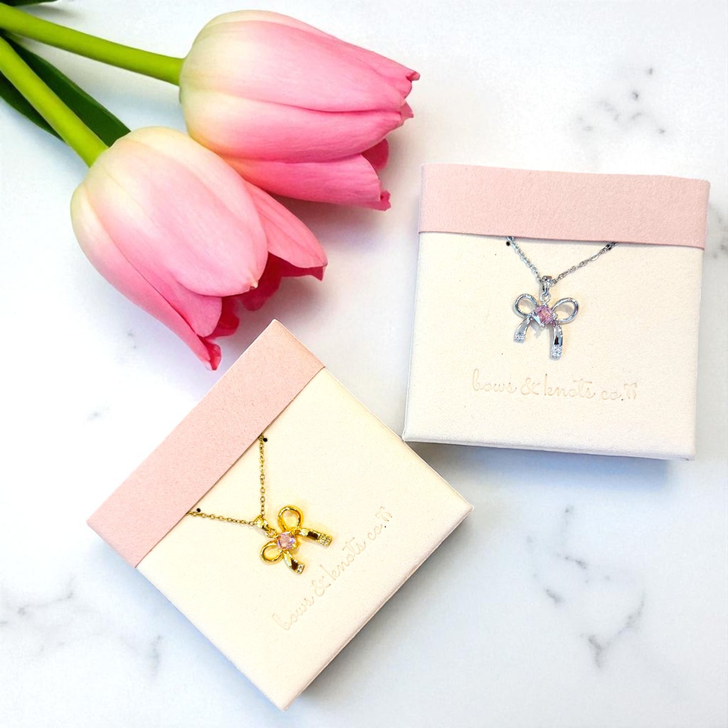 Two bowknot necklaces in display boxes with pink tulips on a marble surface