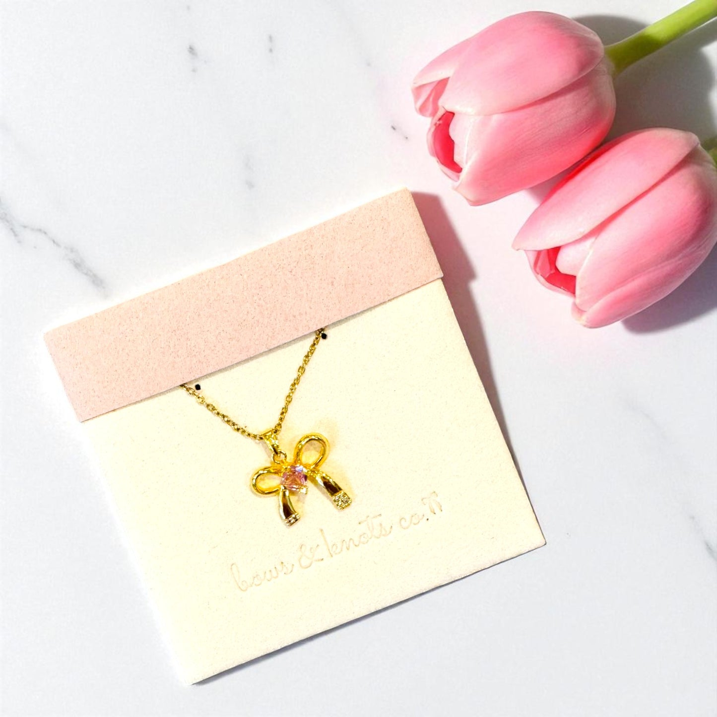 Gold necklace with a bow pendant on a card, next to pink tulips on a light background