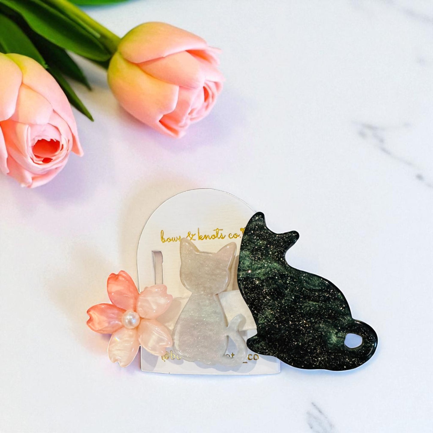 Two cat-shaped hair clips on a white surface with pink flowers and green leaves.