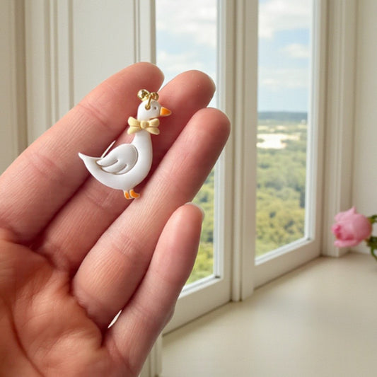 Small white goose earring with a yellow bow tie held in a hand, with a window view in the background.