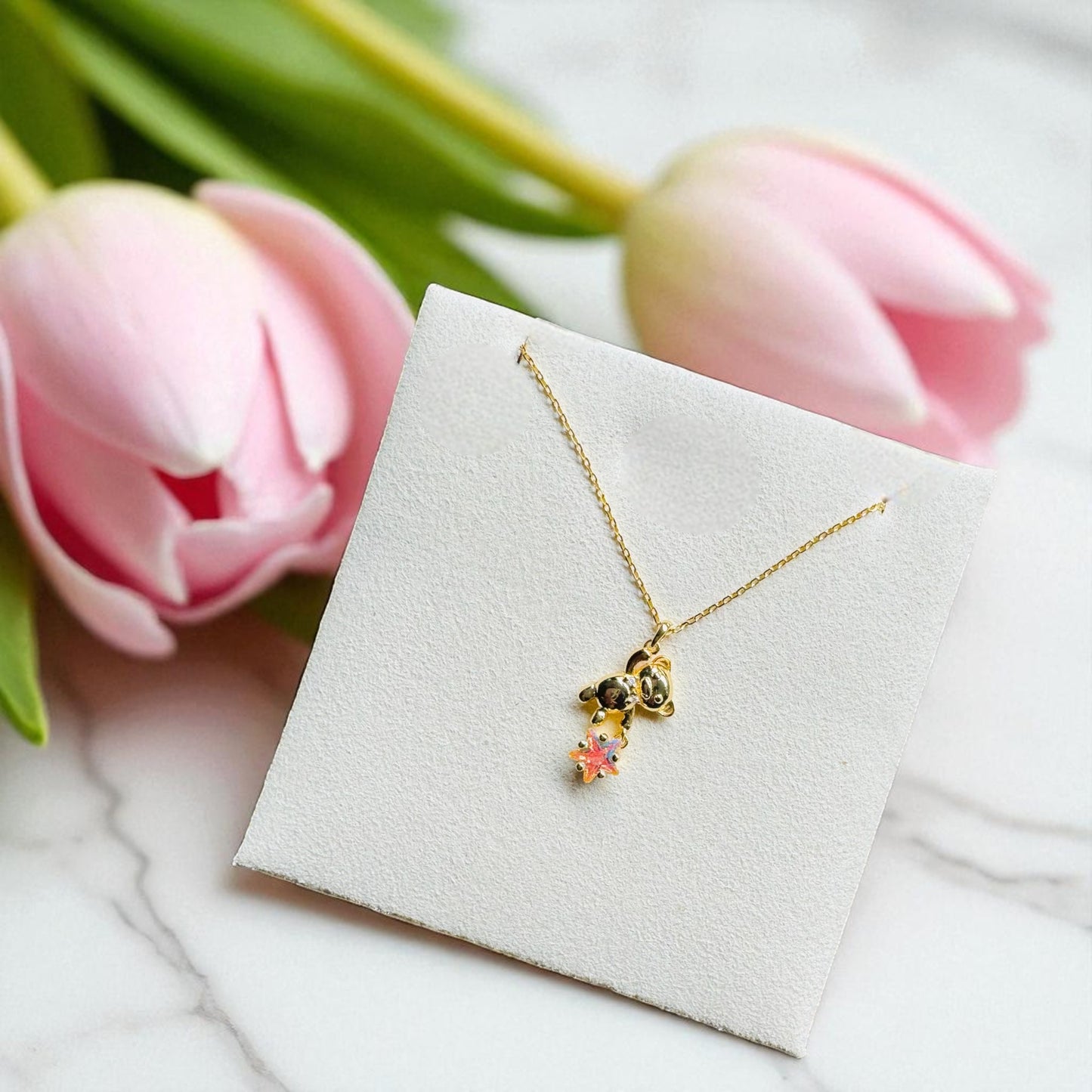Gold necklace with a floral pendant on a white box, surrounded by pink tulips.