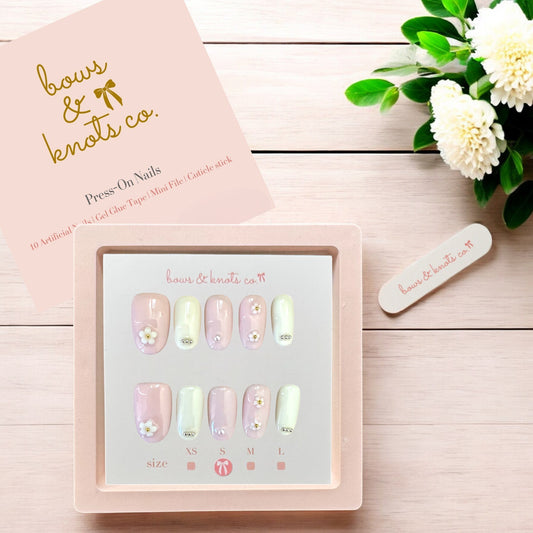 Set of press-on nails with a 'bows & knots co.' box on a wooden surface with flowers.