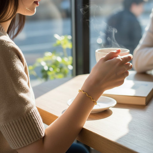 Person holding a steaming cup of coffee wearing a gold wavy chain bracelet from Bows and Knots Co. in a cozy setting with a blurred background