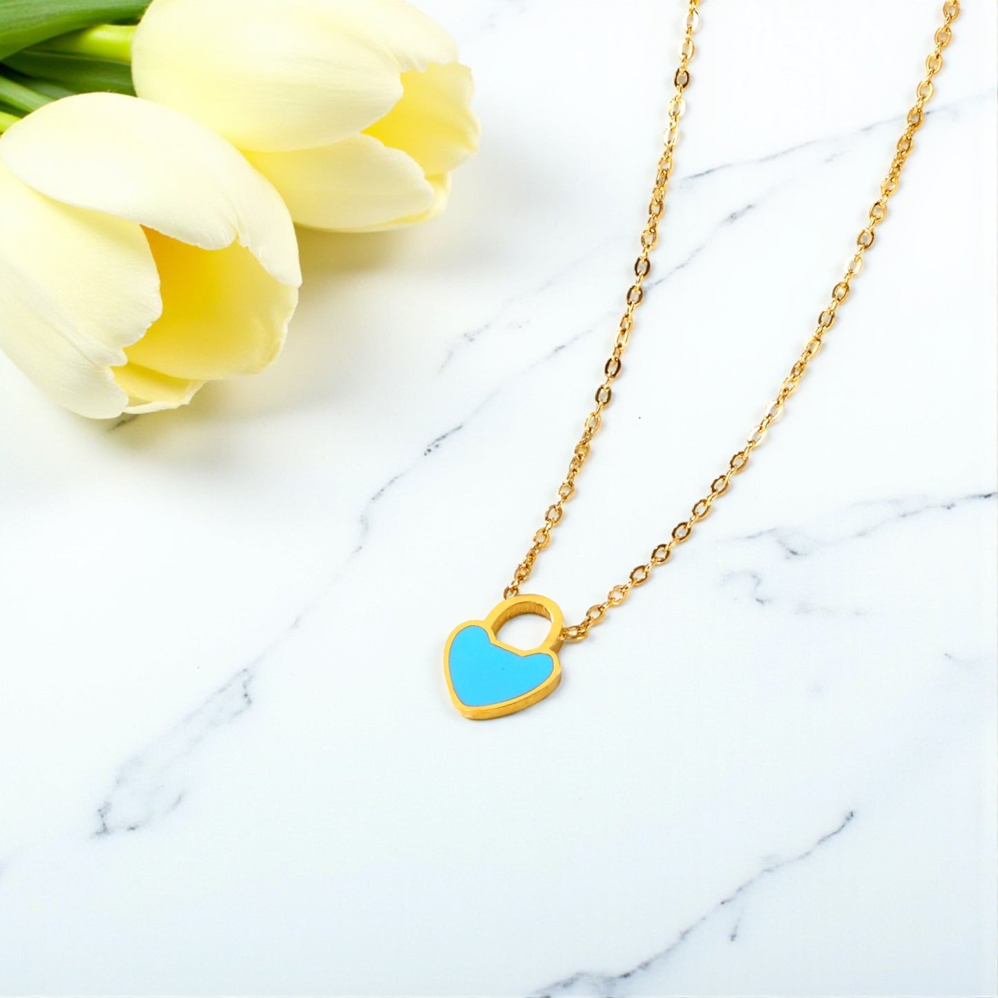 Gold necklace with a blue heart pendant on a marble surface with yellow tulips.