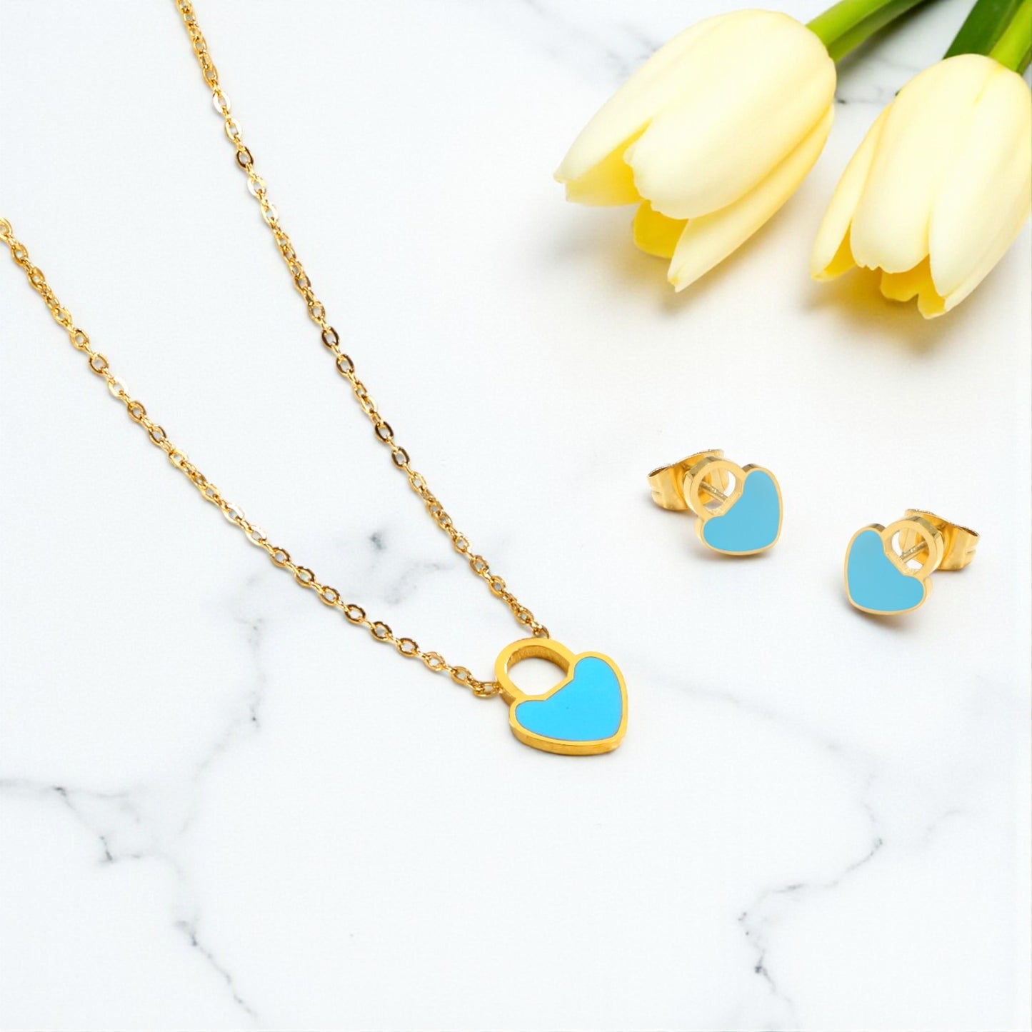 Gold necklace with blue heart pendant and matching earrings on a marble surface with yellow tulips.