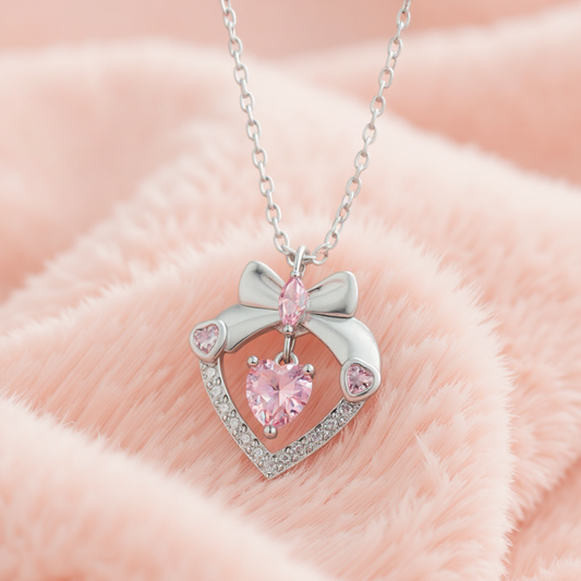 Heart-shaped pendant with pink heart gem and bow design on a chain against a soft pink background