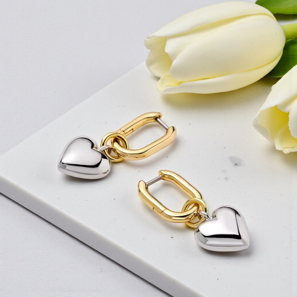 Pair of gold and silver heart-shaped earrings on a marble surface with white tulips.