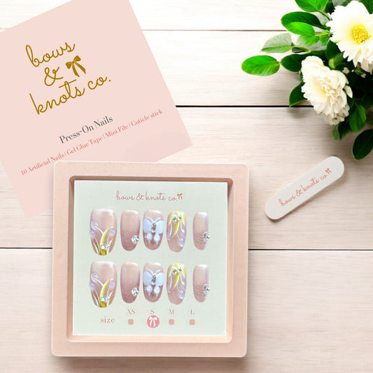 Set of press-on nails with butterfly design in a box labeled 'bows & knots co.' on a wooden surface with flowers.
