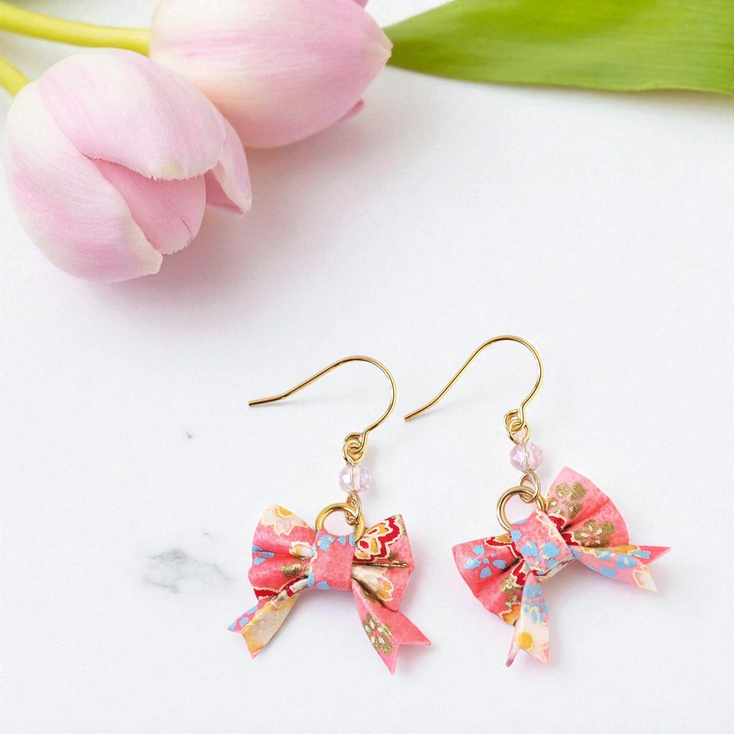 Pair of pink bow-shaped earrings with gold accents on a white background with pink tulips.
