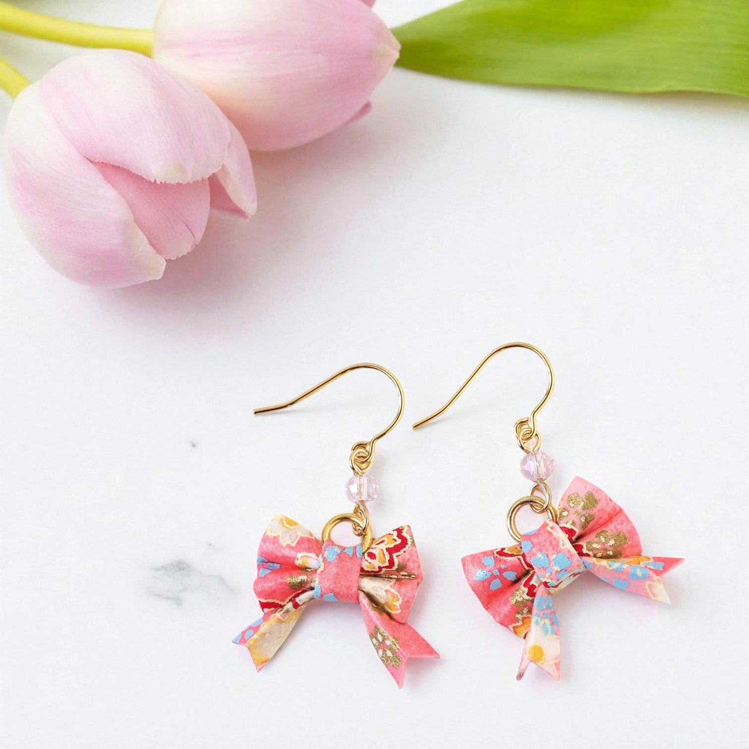 Pair of pink bow-shaped earrings with gold accents on a white background with pink tulips.