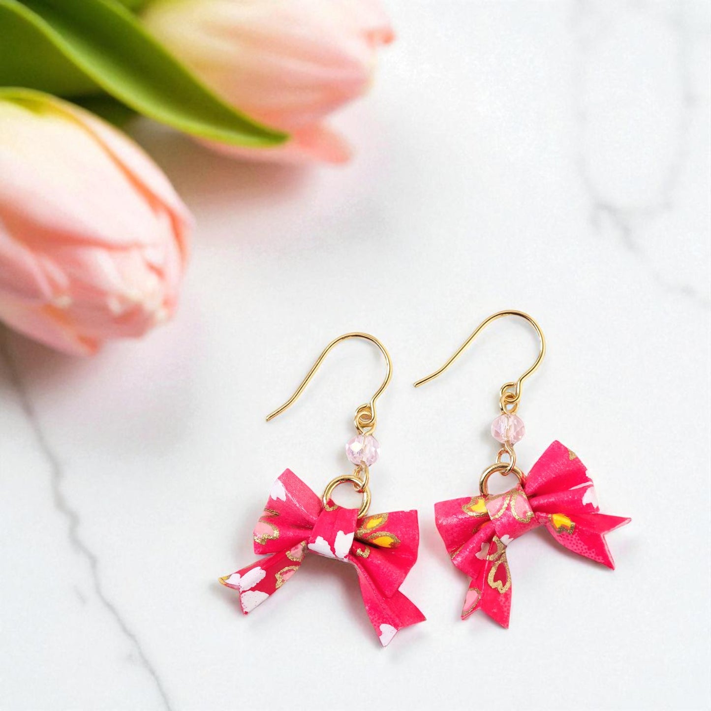 Pink bow-shaped earrings with gold hooks on a white background with pink tulips.