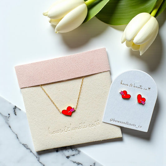 Heart-shaped jewelry set with a necklace and earrings on branded card, surrounded by white tulips.