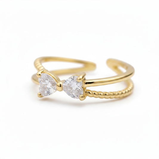 A gold adjustable ring featuring two heart-shaped clear gemstones in a prong setting, with one smooth band and one textured beaded band, shown on a white background.