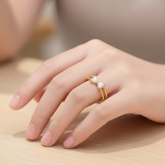 A close-up of a hand wearing a gold ring with two small clear heart-shaped gemstones, resting on a light wooden surface.