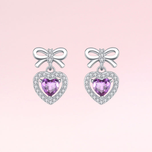 Pair of earrings with heart-shaped purple gemstones and bow designs on a pink background