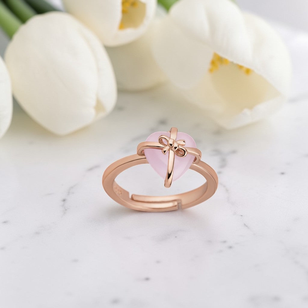 Rose gold ring with a pink heart-shaped gemstone on a marble surface with white tulips in the background.